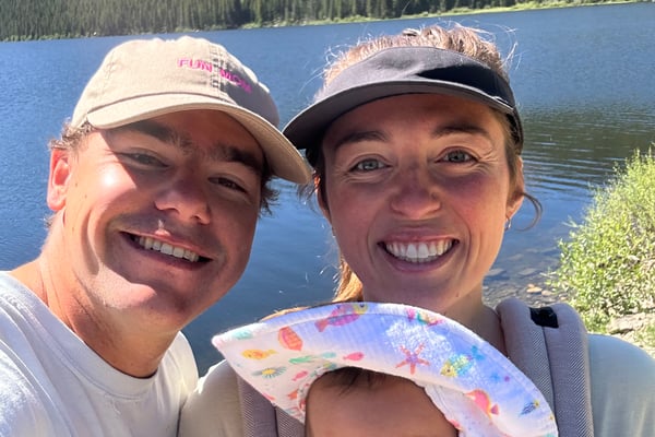 Adult and two young children smiling at camera on a boat with calm water and tree-lined shore in background