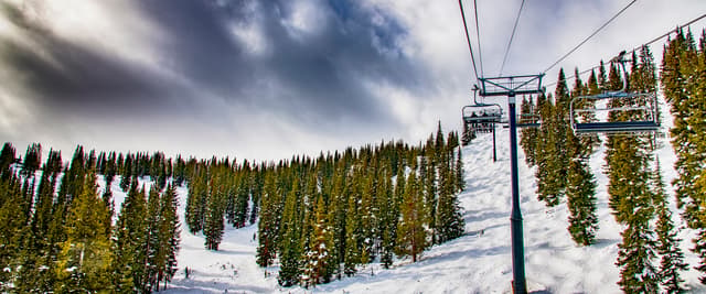 Ski slope with snow-covered terrain, evergreen trees, and ski lift against cloudy sky