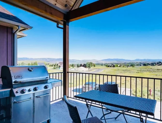 Covered deck with gas grill and dining table overlooking valley, mountains, and distant homes under clear blue sky